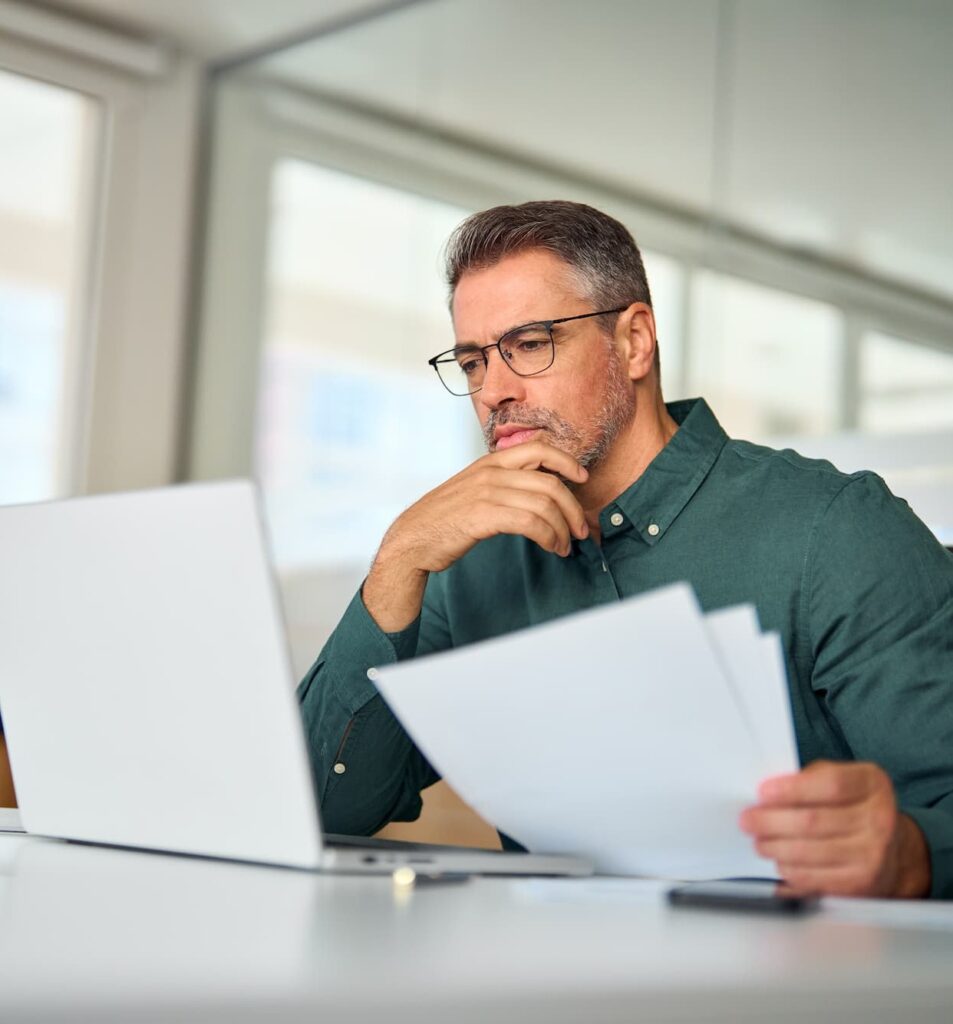Busy middle aged executive, mature male hr manager holding documents using laptop looking at pc in office at desk, thinking over financial data report feeing doubt about market assets investment risk.