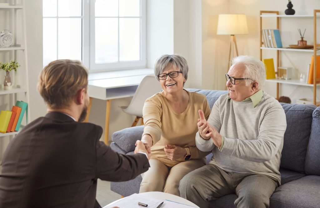 Portrait of happy cheerful senior couple sitting on couch in living room at home shaking hands with male financial advisor reaching agreement in health insurance.
