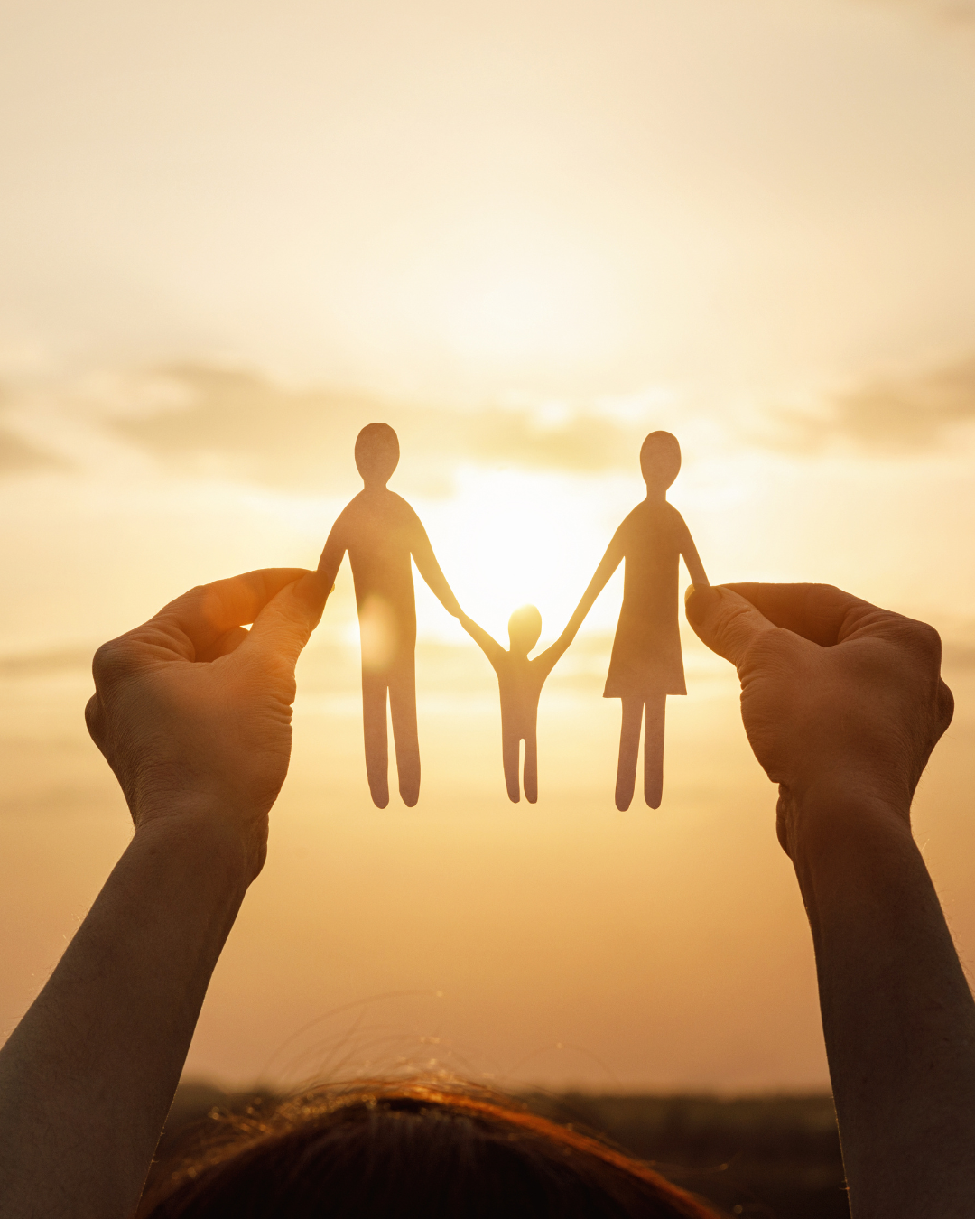 Hands holding a paper cut-out of a family, with two taller figures and a child between them, silhouetted against a warm sunset sky.